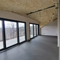 Wnętrze w stanie deweloperskim z wylaną szarą posadzką, oknami balkonowymi z widokiem na drzewa i stropem ocieplonym pianą PUR z wystającymi elementami montażowymi.