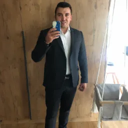 Mężczyzna w garniturze robi selfie przed drewnianą ścianą, obok huśtawki dla dziecka.