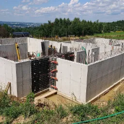 Żelbetowe ściany piwnicy w trakcie budowy, widoczne zbrojenia i szalunki, w tle panorama miasta Rzeszów.