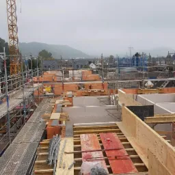 Widok z góry na plac budowy z rusztowaniami, ceglanymi ścianami w trakcie wznoszenia, szalunkami z drewna i żurawiem budowlanym na tle zamglonych wzgórz.