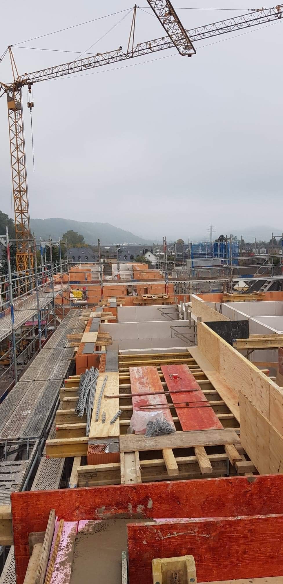 Widok z góry na plac budowy z rusztowaniami, ceglanymi ścianami w trakcie wznoszenia, szalunkami z drewna i żurawiem budowlanym na tle zamglonych wzgórz.