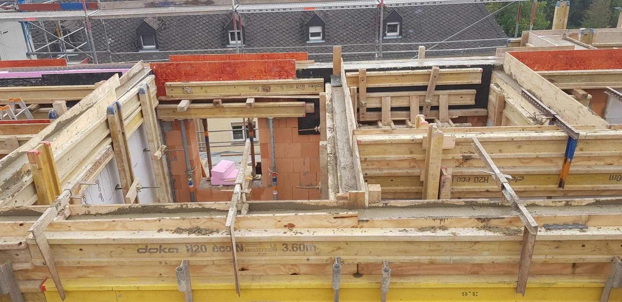 Wysoki kąt widzenia na szalunek stropu z betonu, widoczne drewniane elementy konstrukcyjne, stalowe podpory i fragmenty murowanej ściany z pustaków ceramicznych. W tle dach budynku sąsiedniego...