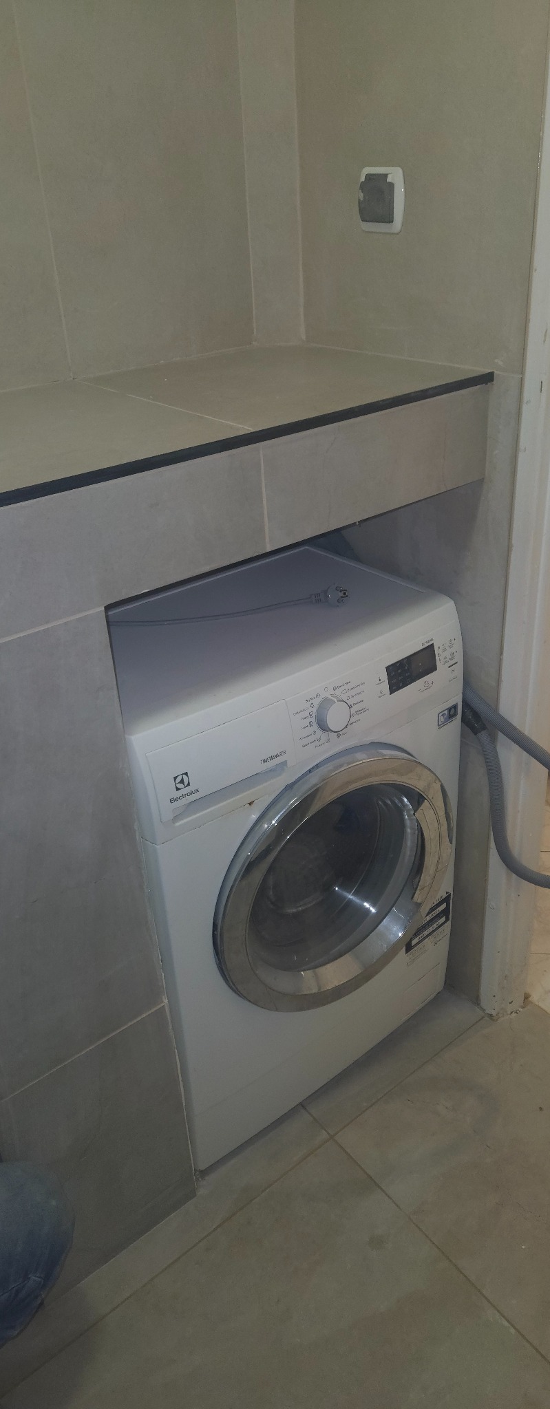 Biała pralka Electrolux w łazience z szarymi płytkami, umieszczona pod blatem. Widoczny fragment podłogi i ściany z gniazdkiem elektrycznym.