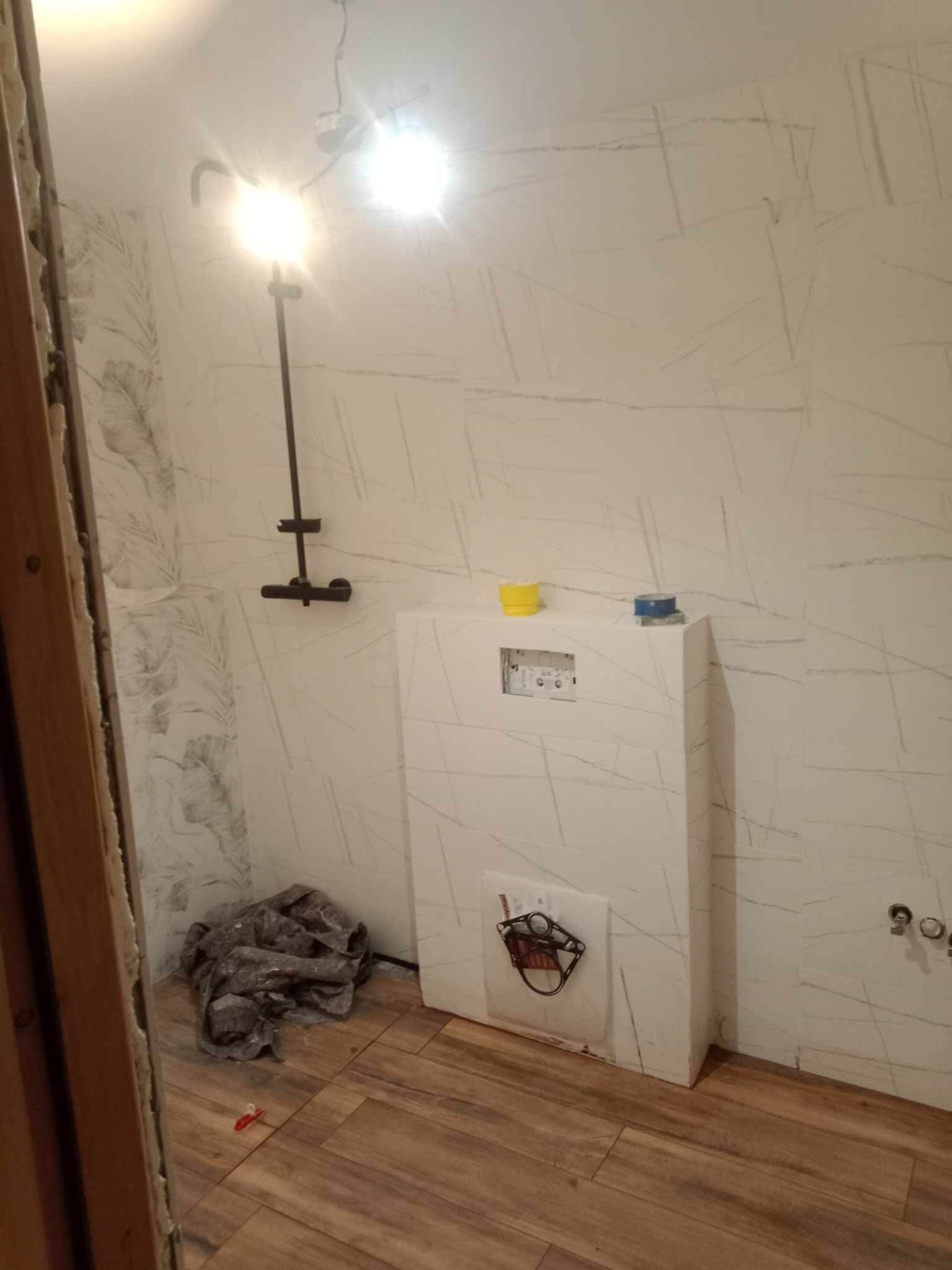 Montaż stelaża podtynkowego WC z widoczną instalacją hydrauliczną i fragmentem ściany z tapetą w liście. Podłoga wyłożona panelami imitującymi drewno.