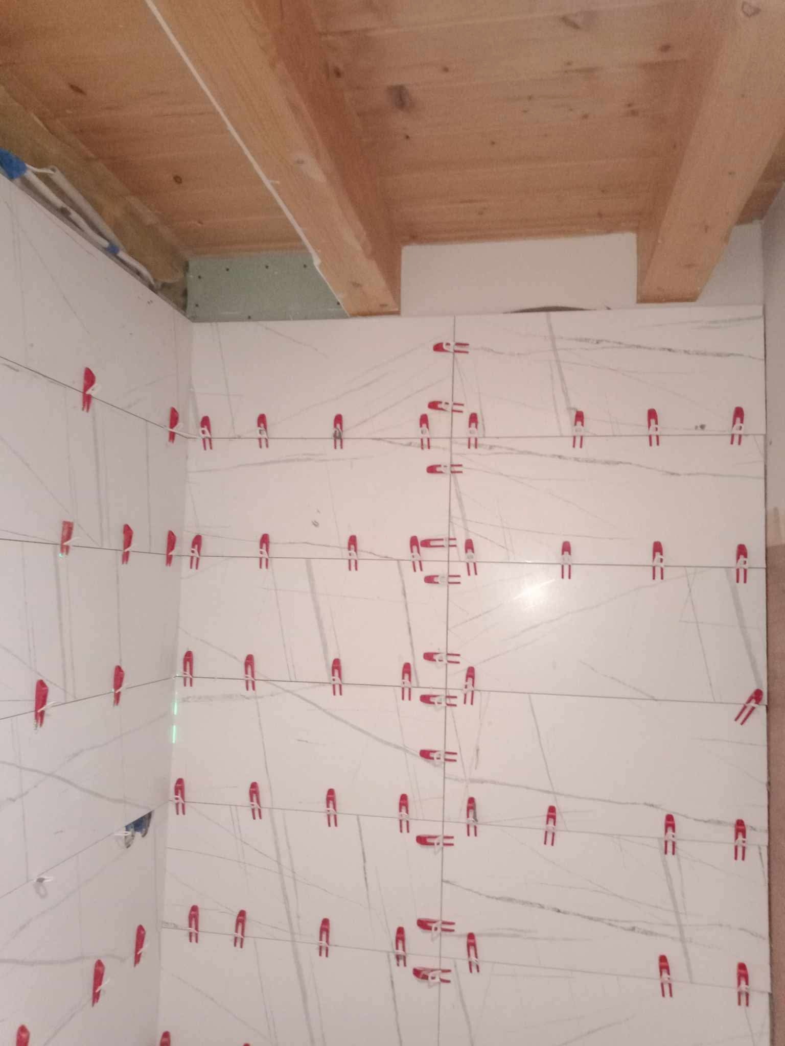 Ściana w trakcie układania białych płytek z szarymi żyłkami, z użyciem czerwonych klipsów poziomujących i lasera krzyżowego. Widoczny fragment drewnianego sufitu.