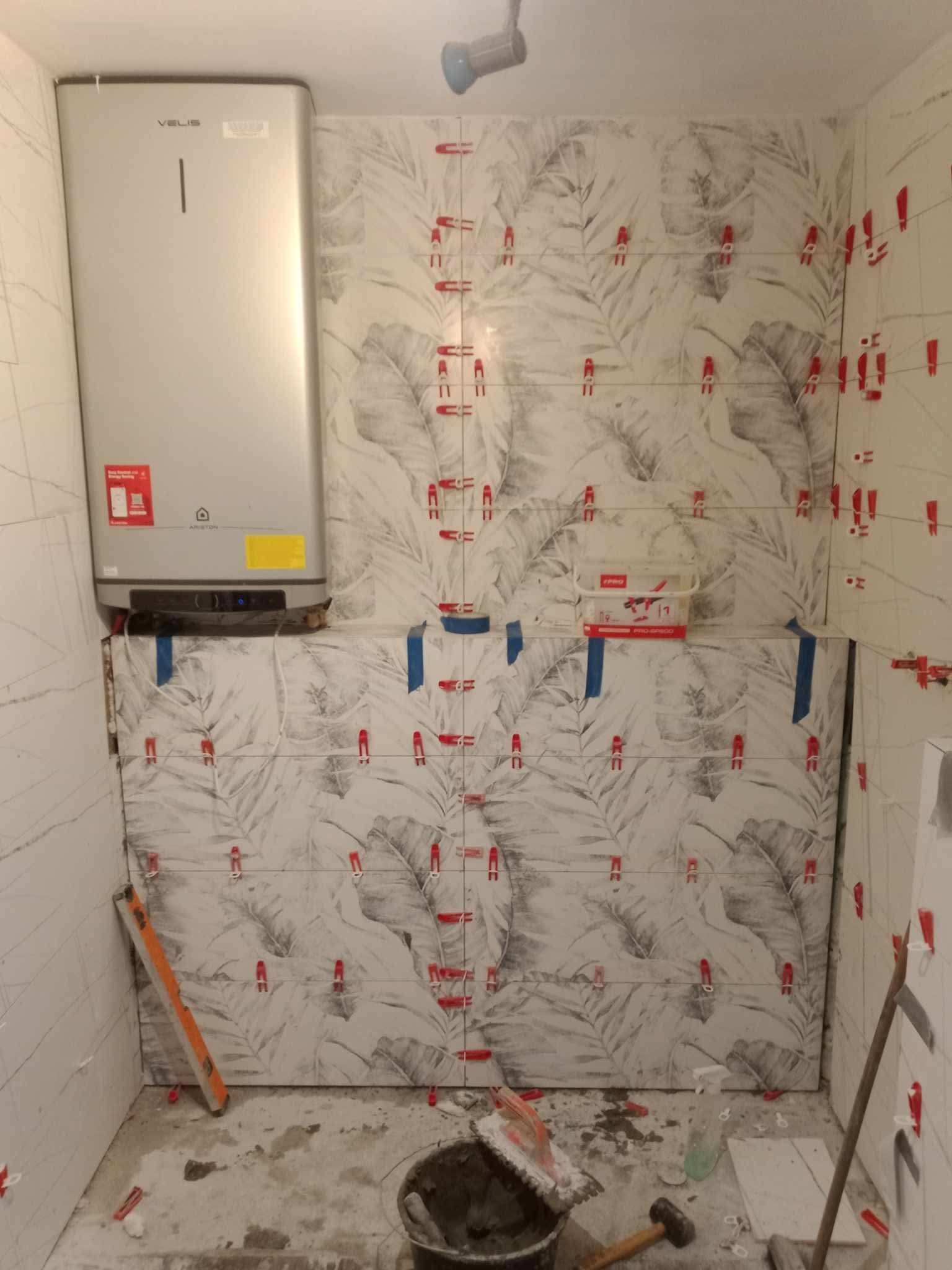 Układanie płytek z motywem liści w łazience, widoczne narzędzia i poziomica. Częściowo wykończona ściana, grzejnik elektryczny na ścianie, czerwone klipsy poziomujące płytki.