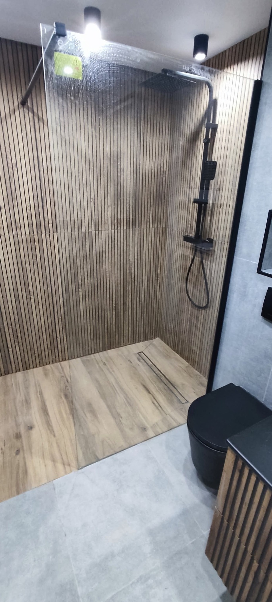 Nowoczesna kabina prysznicowa walk-in z drewnopodobnymi panelami ściennymi i czarną armaturą. Minimalistyczny design, widoczny odpływ liniowy w podłodze.