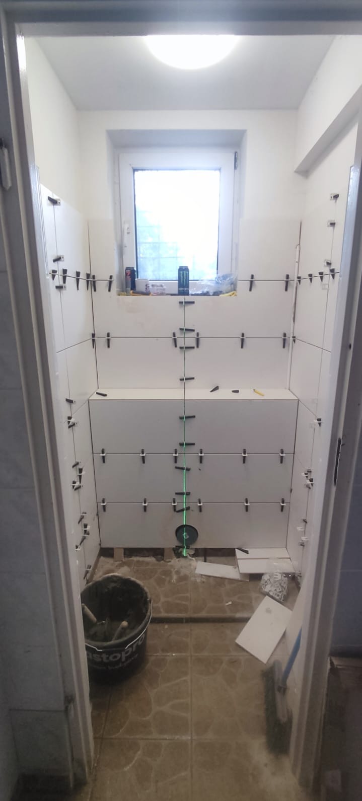 Białe płytki układane w łazience, widoczne poziomice laserowe i krzyżyki dystansowe. Prace wykończeniowe w trakcie realizacji, widok z perspektywy wejścia.