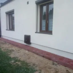 Świeżo wykończony budynek z białymi ścianami, brązowymi oknami z roletami i obrzeżem z czerwonej kostki brukowej wzdłuż fundamentów, widoczne trawnik i przygotowany teren pod nasadzenia.