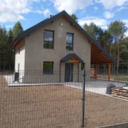 Dwukondygnacyjny dom jednorodzinny z szarą elewacją i ciemną stolarką okienną, widoczny zza ogrodzenia panelowego na tle lasu, z zadaszeniem nad wejściem i drewnianą konstrukcją z tyłu.