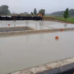 Świeżo wylana płyta fundamentowa z wystającymi prętami zbrojeniowymi, widoczne elementy szalunku i izolacji, w tle zielony krajobraz.