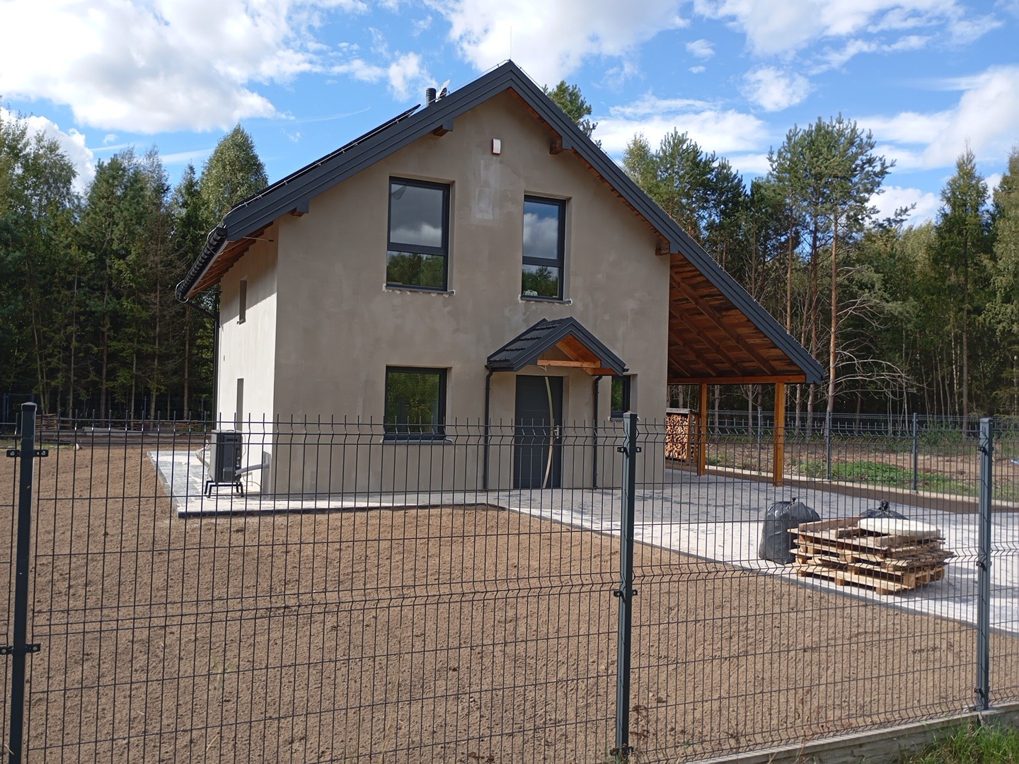 Dwukondygnacyjny dom jednorodzinny z szarą elewacją i ciemną stolarką okienną, widoczny zza ogrodzenia panelowego na tle lasu, z zadaszeniem nad wejściem i drewnianą konstrukcją z tyłu.