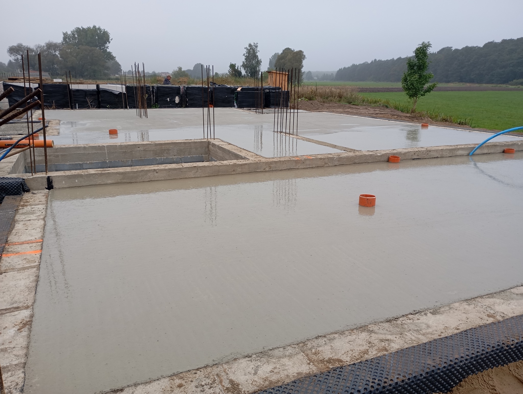 Świeżo wylana płyta fundamentowa z wystającymi prętami zbrojeniowymi, widoczne elementy szalunku i izolacji, w tle zielony krajobraz.