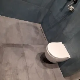 Nowoczesna łazienka z szarymi płytkami imitującymi beton, kabiną prysznicową typu walk-in i podwieszaną toaletą z deską wolnoopadającą.