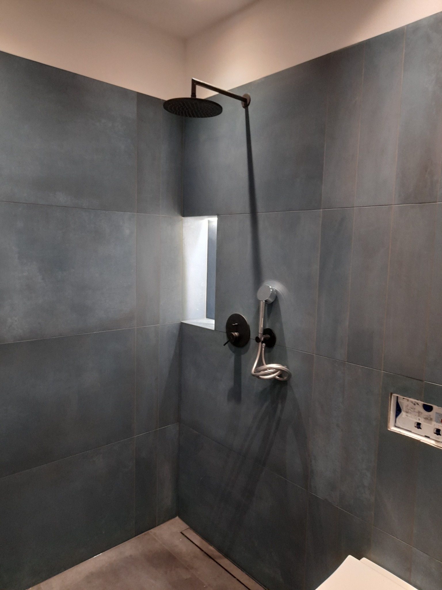 Nowoczesna kabina prysznicowa z grafitowymi, matowymi płytkami wielkoformatowymi, deszczownicą w kolorze czarnym i wnęką z oświetleniem.