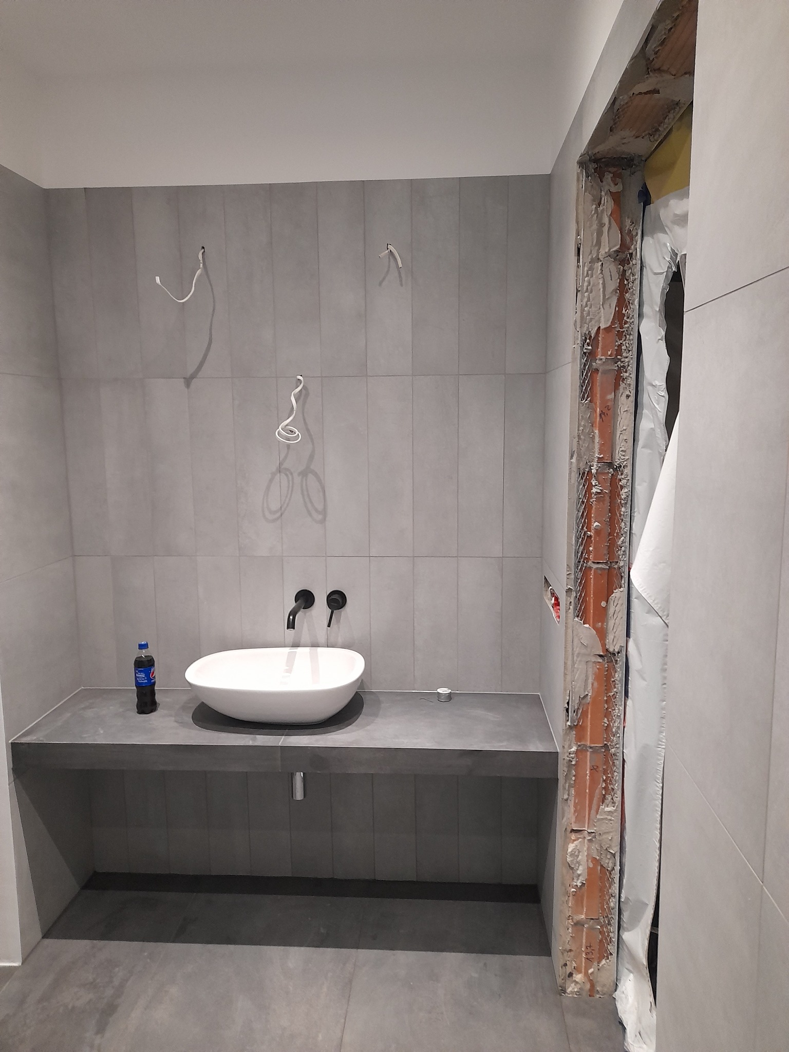 Szara łazienka w trakcie remontu: ułożone płytki, biała umywalka nablatowa z czarną armaturą, widoczne przewody elektryczne i niezakończone obramowanie drzwi z odsłoniętą cegłą.