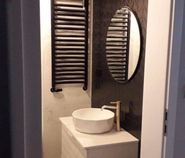 Mała łazienka z owalnym lustrem na czarnej ścianie z geometrycznym wzorem, białą umywalką nablatową z złotą baterią i czarnym grzejnikiem drabinkowym.