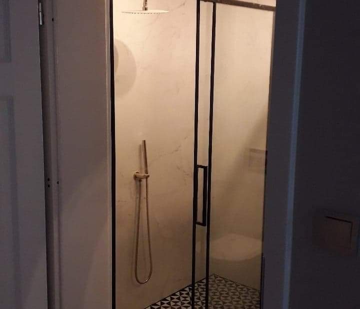 Kabina prysznicowa z czarnymi profilami, deszczownicą i złotą słuchawką na tle marmurowej ściany, wzorzysta podłoga.