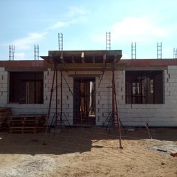 Mał-Bud Kinga Małecka - Wznoszenie ścian parterowego budynku z betonu komórkowego, widoczne szalunki nad otworami okiennymi i zbrojenie wieńca, palety z bloczkami obok zaparkowanego samochodu.