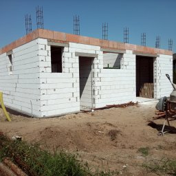 Mał-Bud Kinga Małecka - Wznoszenie ścian parterowego domu z białych bloczków, widoczne otwory okienne i drzwiowe, wystające pręty zbrojeniowe, obok stoi brudna betoniarka, w tle zabudowa jednorodzinna.