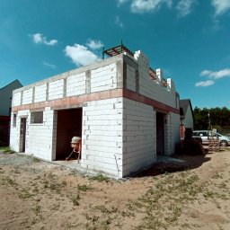 Mał-Bud Kinga Małecka - Surowy stan budynku w trakcie budowy z białych bloczków, z widocznymi czerwonymi elementami ceglanymi, na tle błękitnego nieba z chmurami. Widoczna betoniarka w środku budynku i palety...