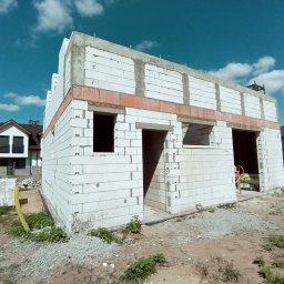 Mał-Bud Kinga Małecka - Niedokończony budynek z białych bloczków, widoczne otwory okienne i drzwiowe, w tle inne domy i niebieskie niebo z chmurami.