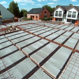 Mał-Bud Kinga Małecka - Wieniec stropowy w trakcie budowy domu murowanego, widok z góry na bloczki betonowe i zbrojenie stalowe, w tle zabudowa jednorodzinna i błękitne niebo z chmurami.