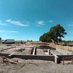 Wylane fundamenty pod budynek z widocznymi zbrojeniami, ujęcie w słoneczny dzień z perspektywy szerokokątnej, na horyzoncie widoczny krajobraz polny i biały samochód dostawczy.