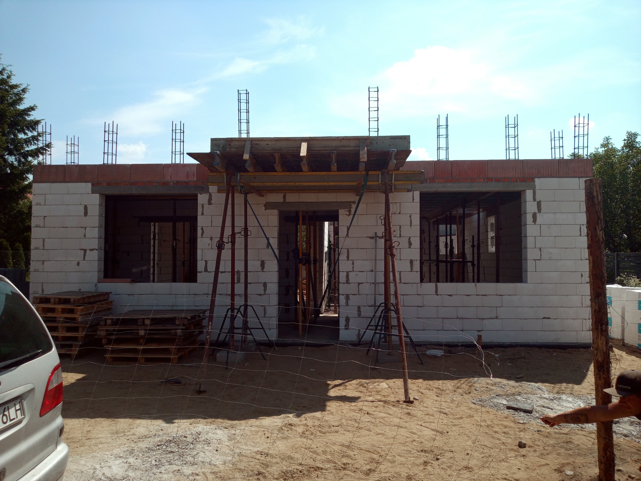 Wznoszenie ścian parterowego budynku z betonu komórkowego, widoczne szalunki nad otworami okiennymi i zbrojenie wieńca, palety z bloczkami obok zaparkowanego samochodu.