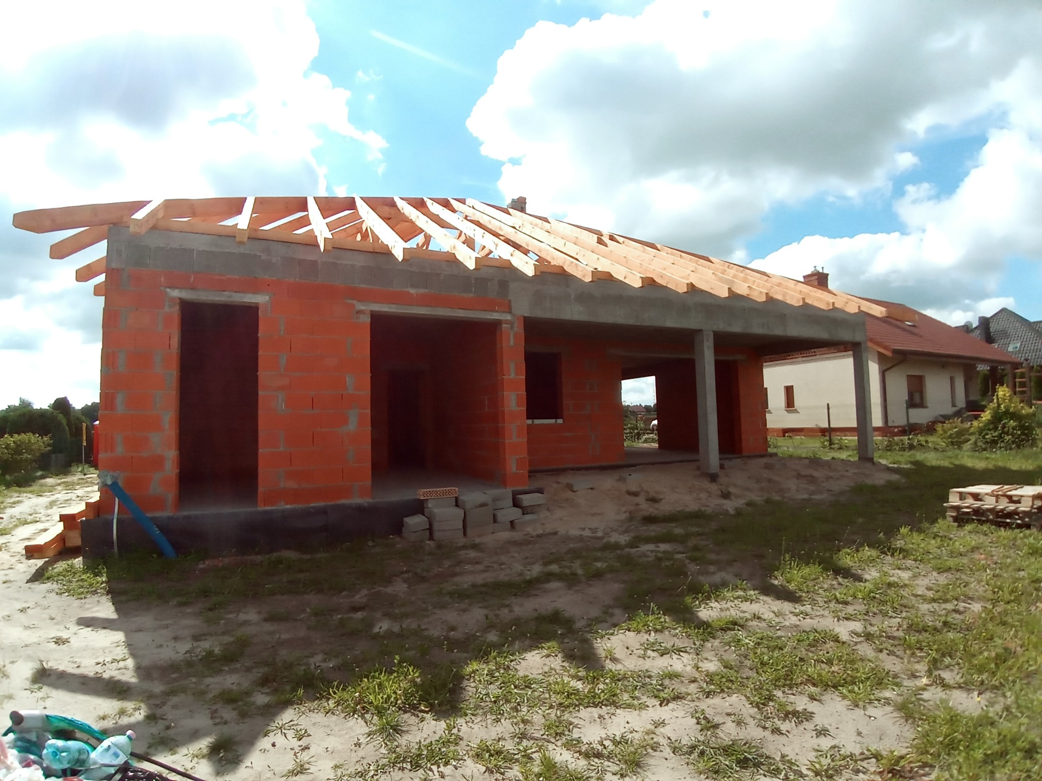 Niedokończony budynek z czerwonej cegły z drewnianą konstrukcją dachu, widok z zewnątrz, na tle błękitnego nieba z chmurami.