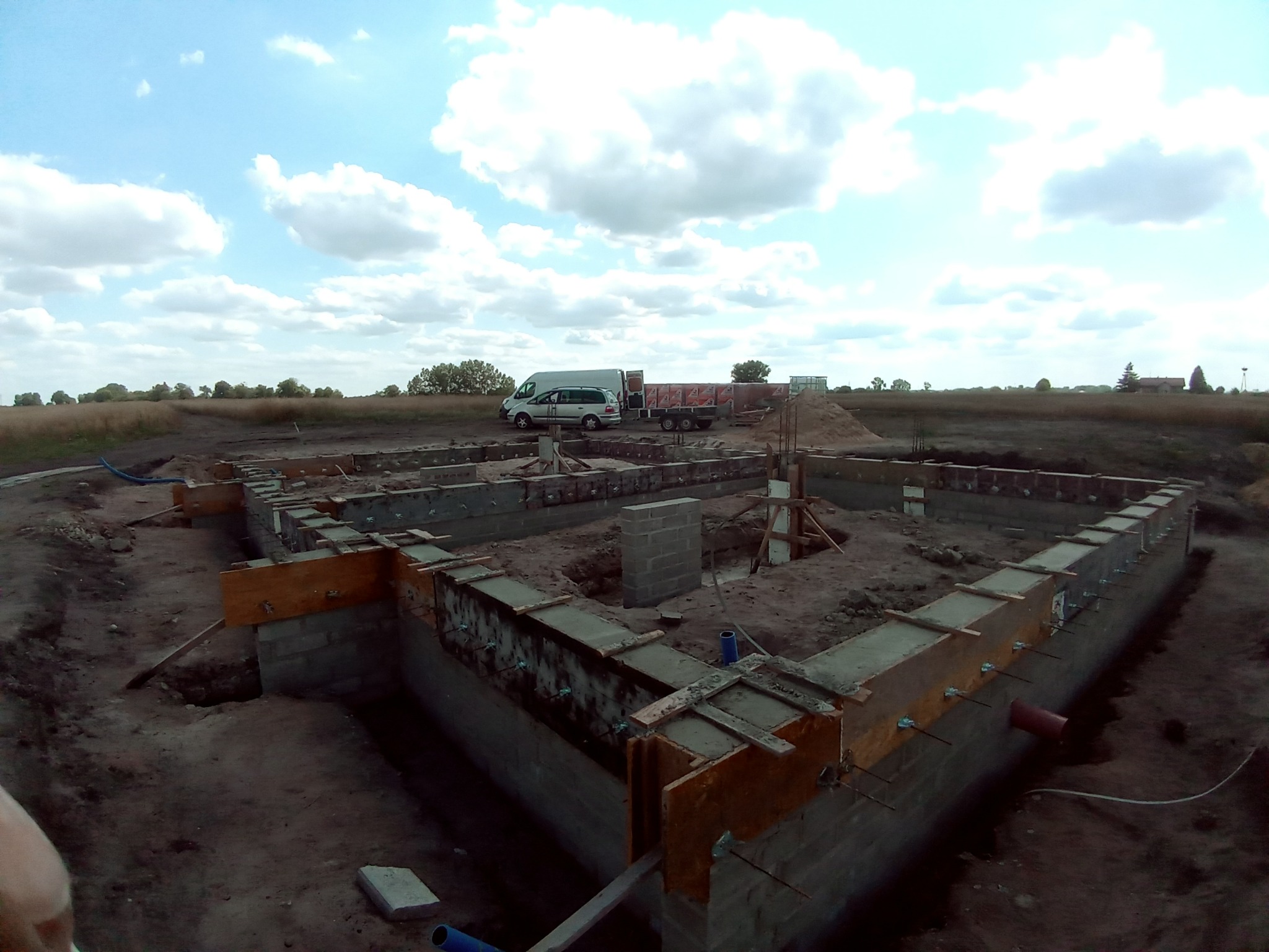 Wykopane i przygotowane fundamenty pod budowę domu z widocznym szalunkiem, zbrojeniami, częściowo wylane betonem, na tle krajobrazu z samochodem dostawczym i przyczepą