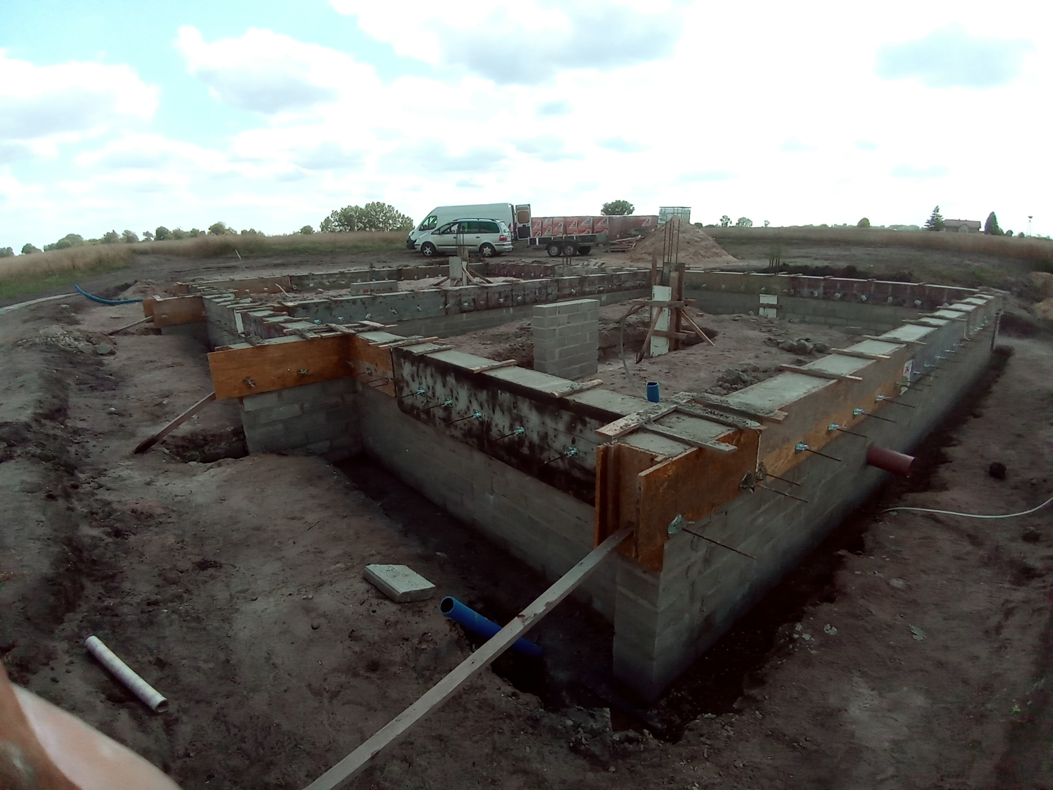 Wykop pod fundamenty z częściowo wylanymi ścianami, drewnianym szalunkiem i wystającymi prętami zbrojeniowymi, na tle krajobrazu z samochodem dostawczym i przyczepą.