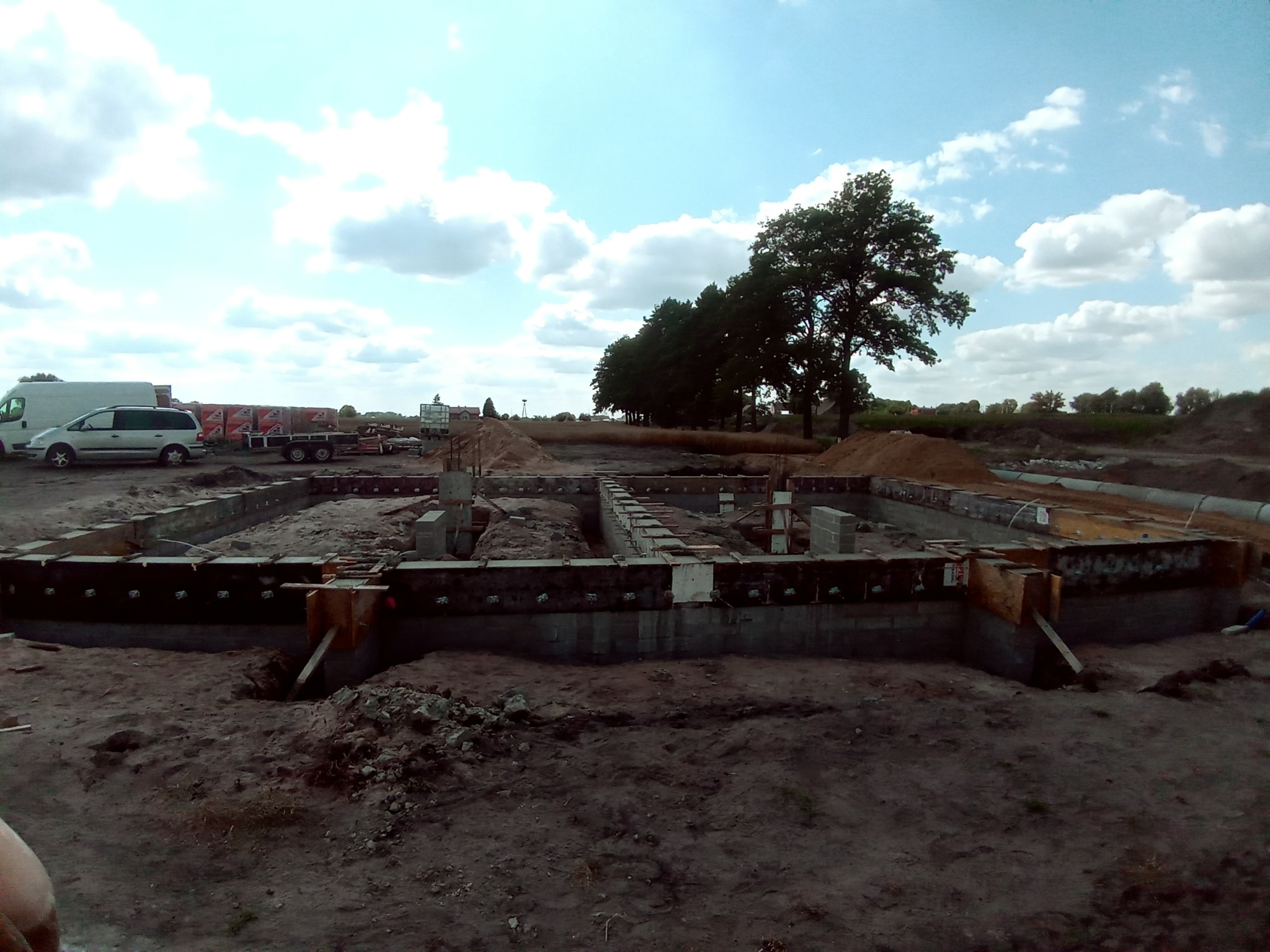 Zalane fundamenty domu jednorodzinnego z widocznym szalunkiem i zbrojeniem, na tle plac budowy z zaparkowanymi pojazdami i drzewami.