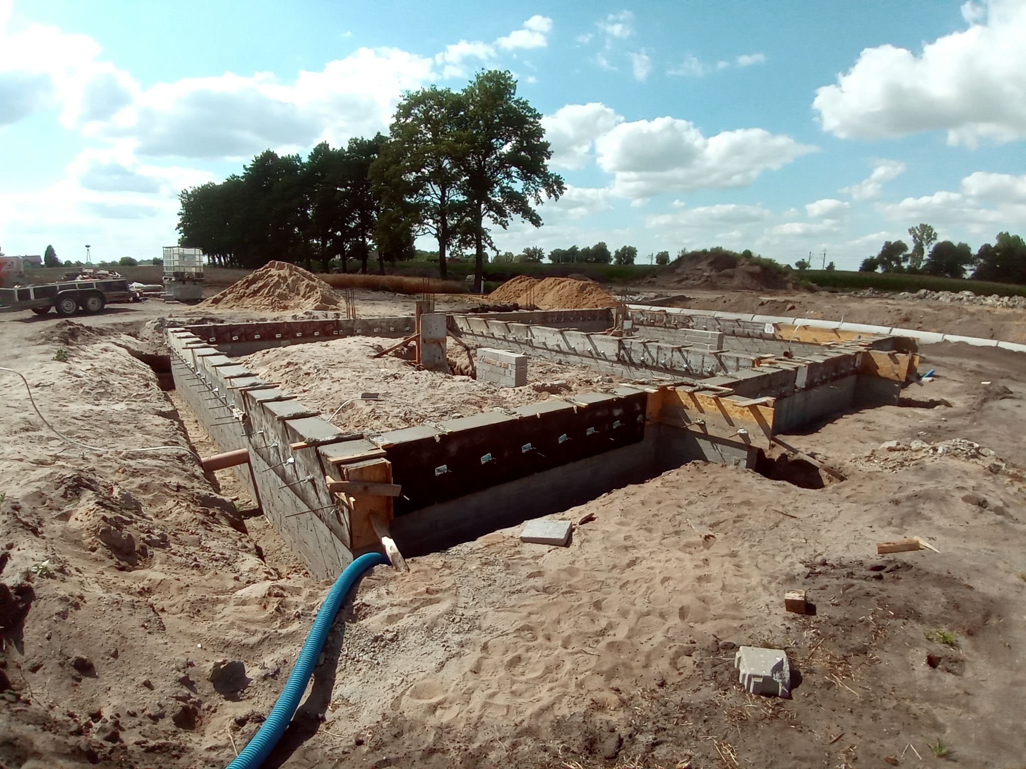 Wykopane fundamenty z drewnianym szalunkiem, częściowo zasypane piaskiem, widoczne rury kanalizacyjne i traktor w tle, ujęcie z góry.