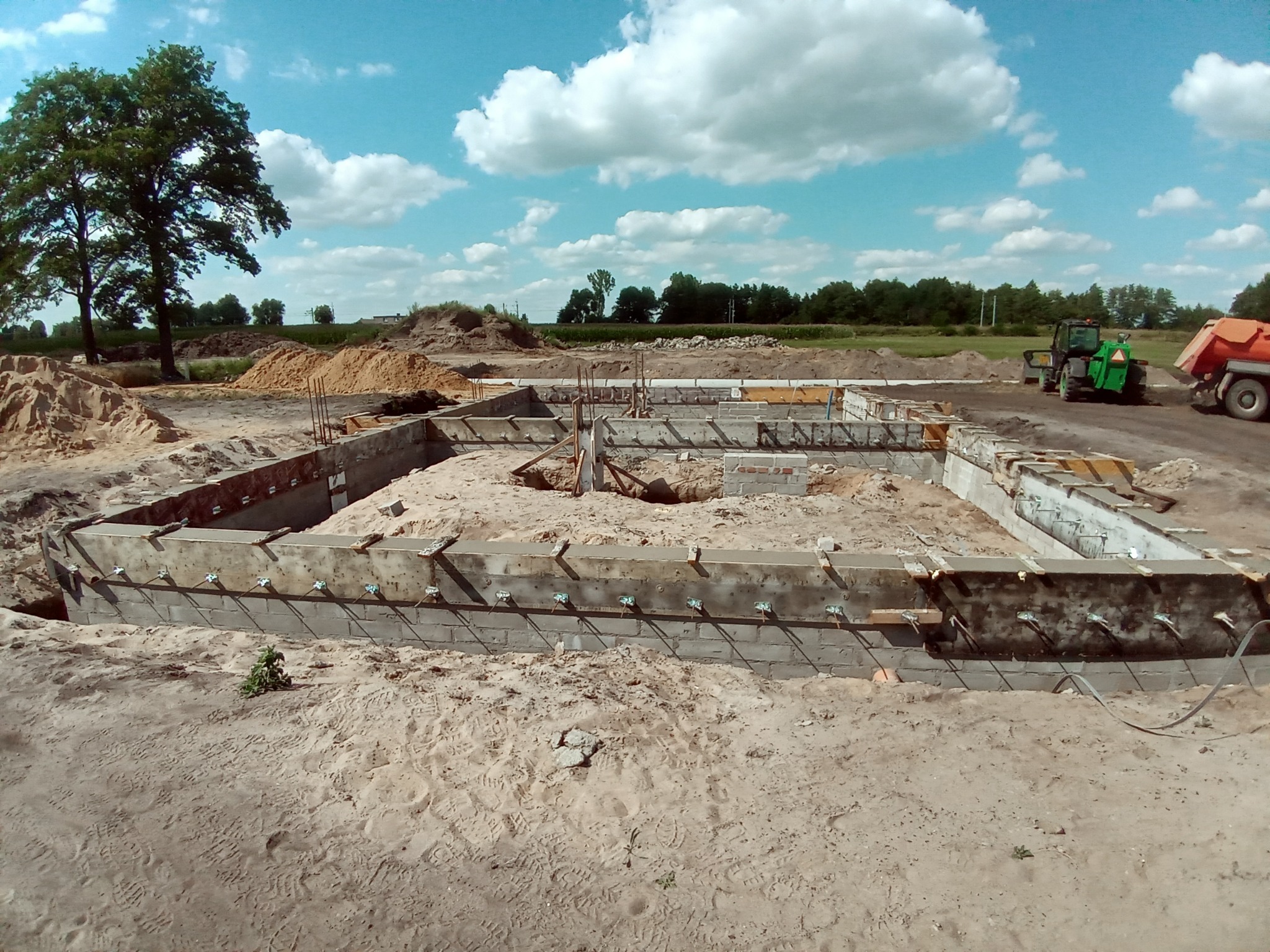 Wykop pod fundamenty domu jednorodzinnego z deskowaniem i zbrojeniami, na piaszczystym terenie, w tle sprzęt budowlany i drzewa.