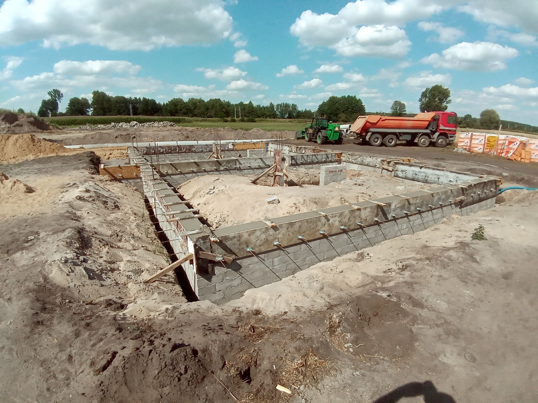 Wykop pod fundamenty domu z częściowo wylanym betonem, widoczne szalunki, zbrojenie, koparka i ciężarówka w tle.