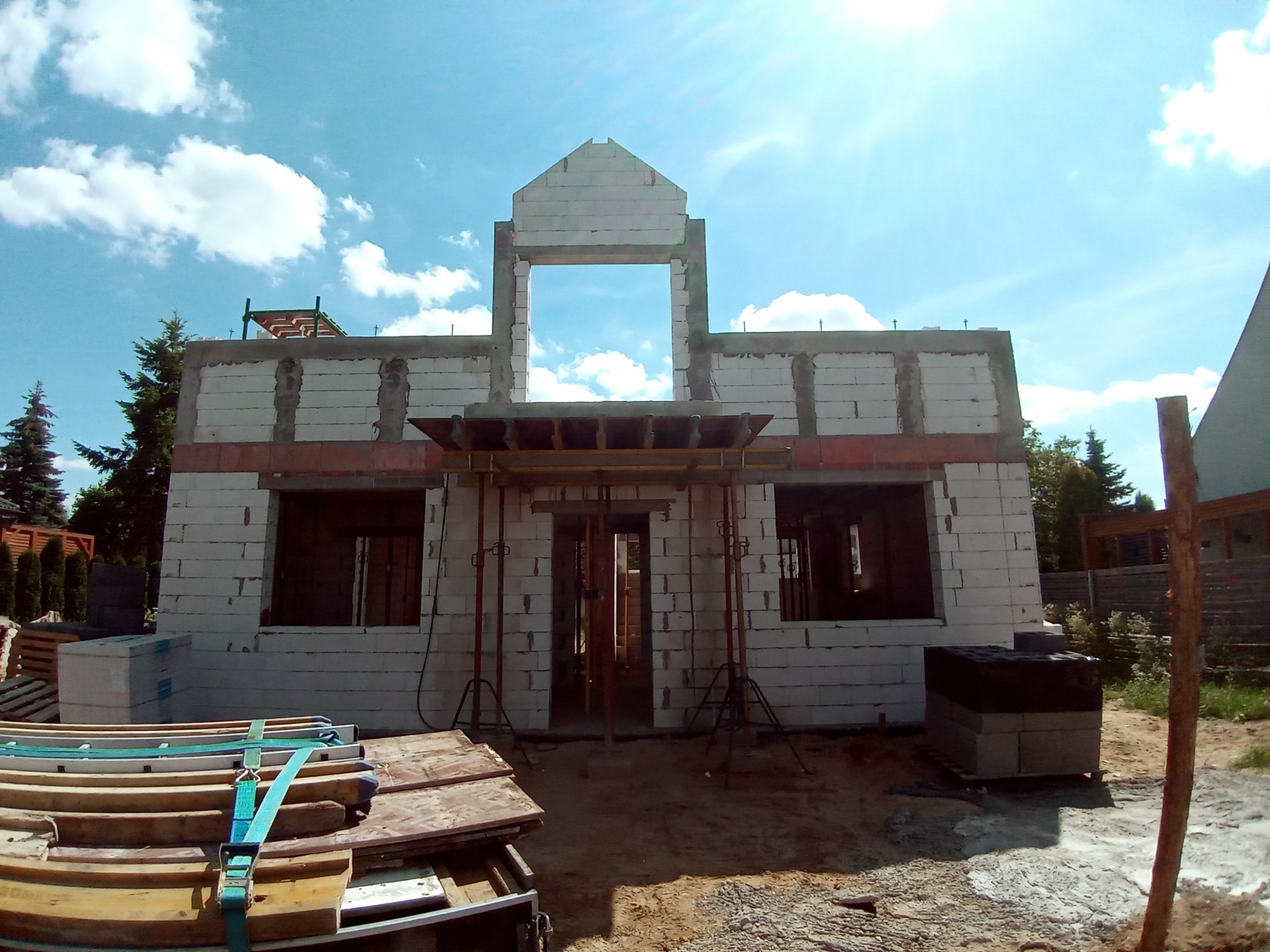 Surowy stan domu z białych bloczków, widoczne otwory okienne i drzwiowe, podparte szalunki, błękitne niebo w tle.