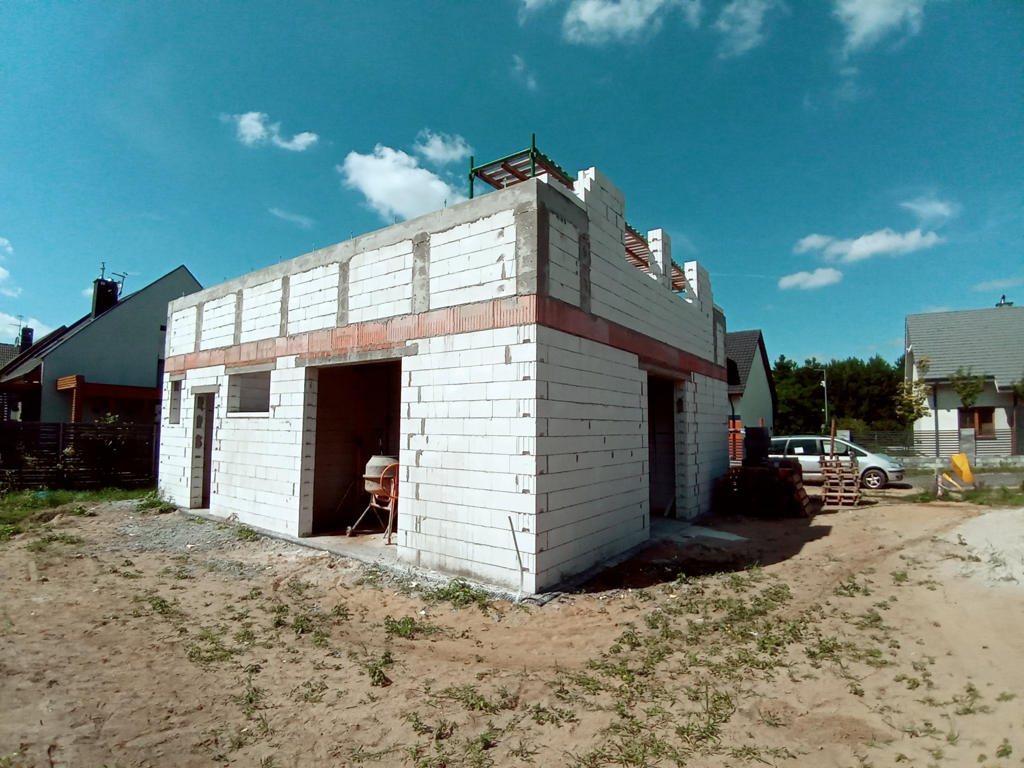 Surowy stan budynku w trakcie budowy z białych bloczków, z widocznymi czerwonymi elementami ceglanymi, na tle błękitnego nieba z chmurami. Widoczna betoniarka w środku budynku i palety...