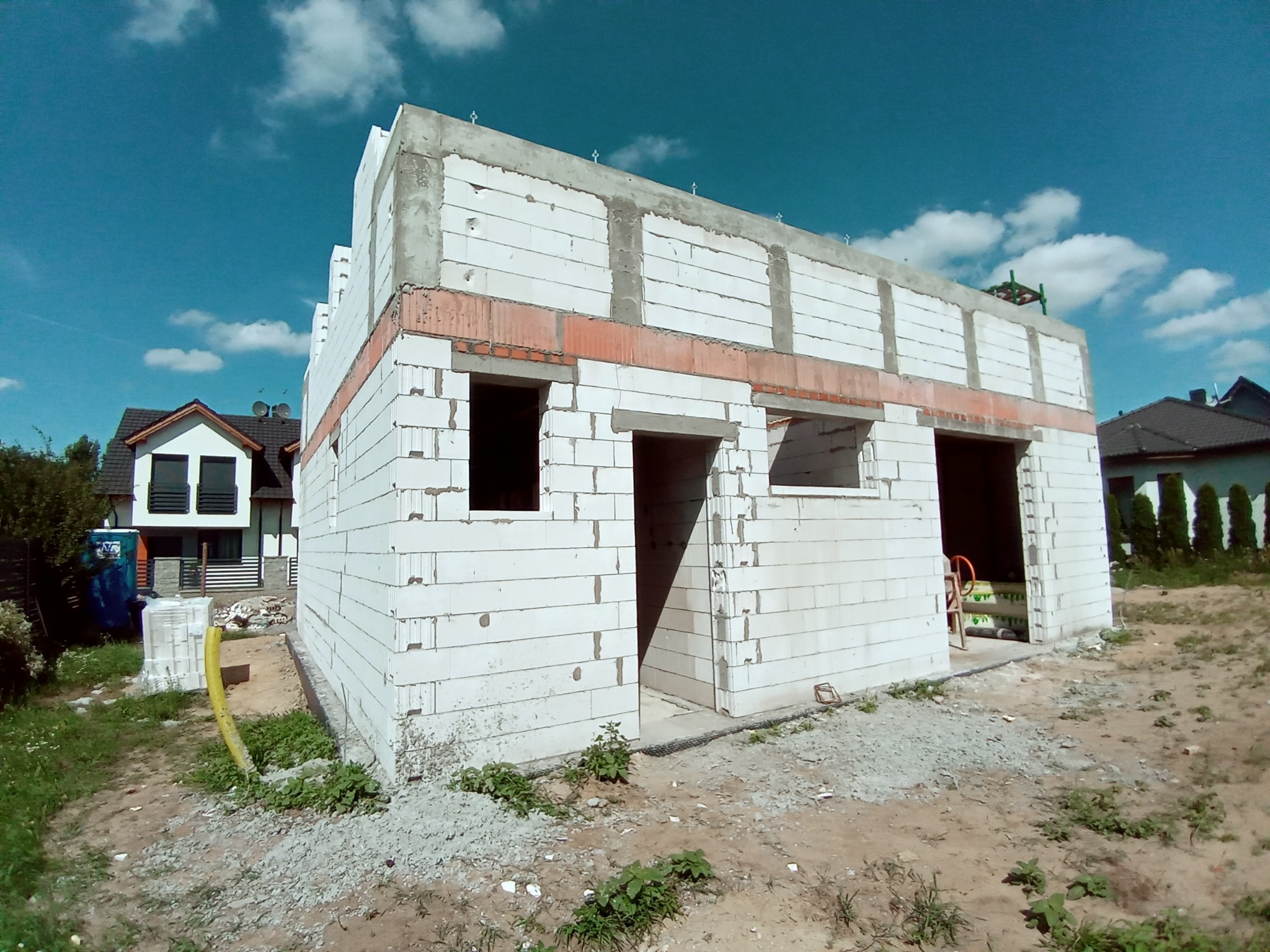 Niedokończony budynek z białych bloczków, widoczne otwory okienne i drzwiowe, w tle inne domy i niebieskie niebo z chmurami.