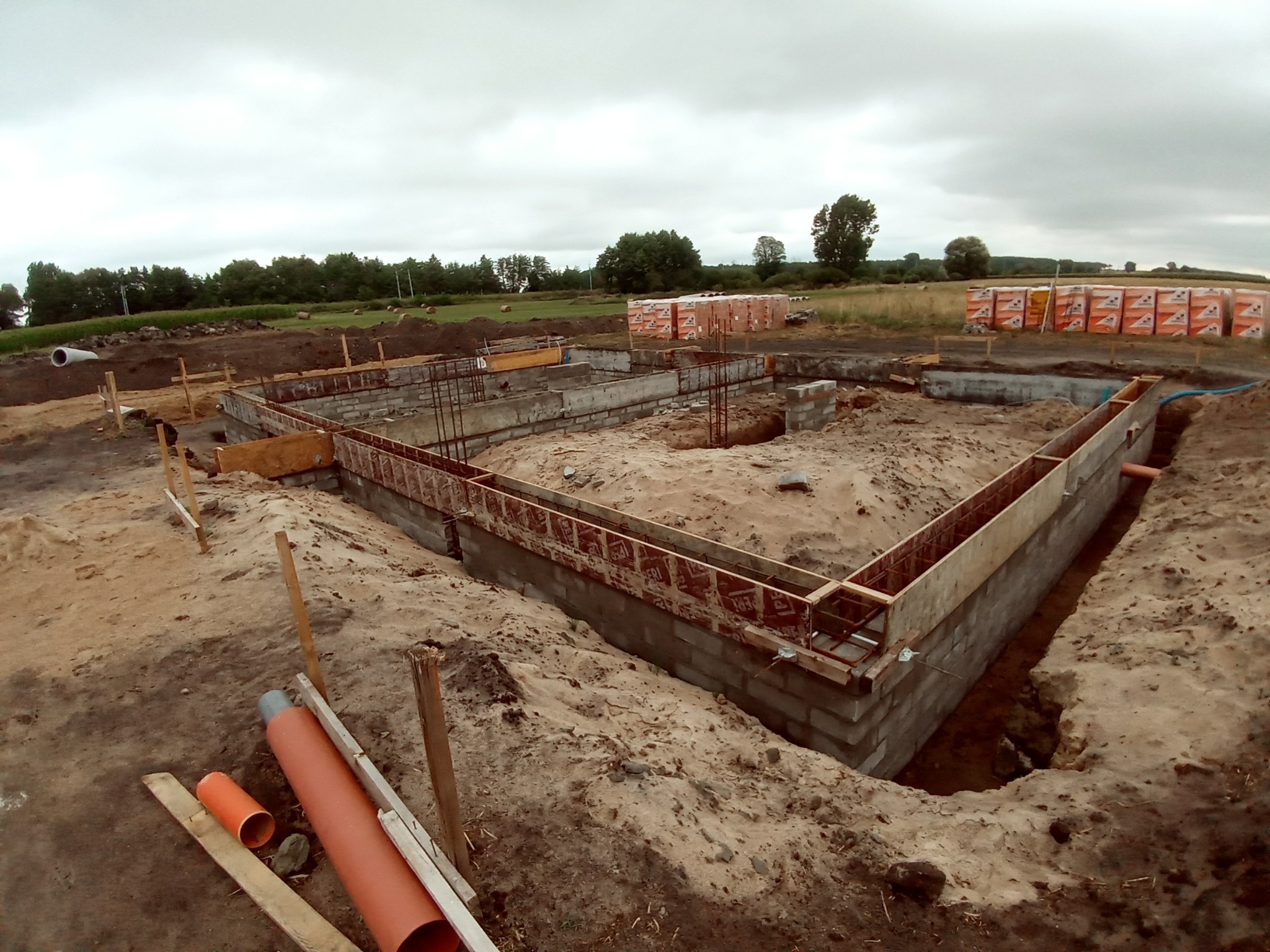 Wykop pod fundamenty domu jednorodzinnego z widocznymi bloczkami betonowymi, zbrojeniami i szalunkami drewnianymi, w tle ułożone palety z cegłami.