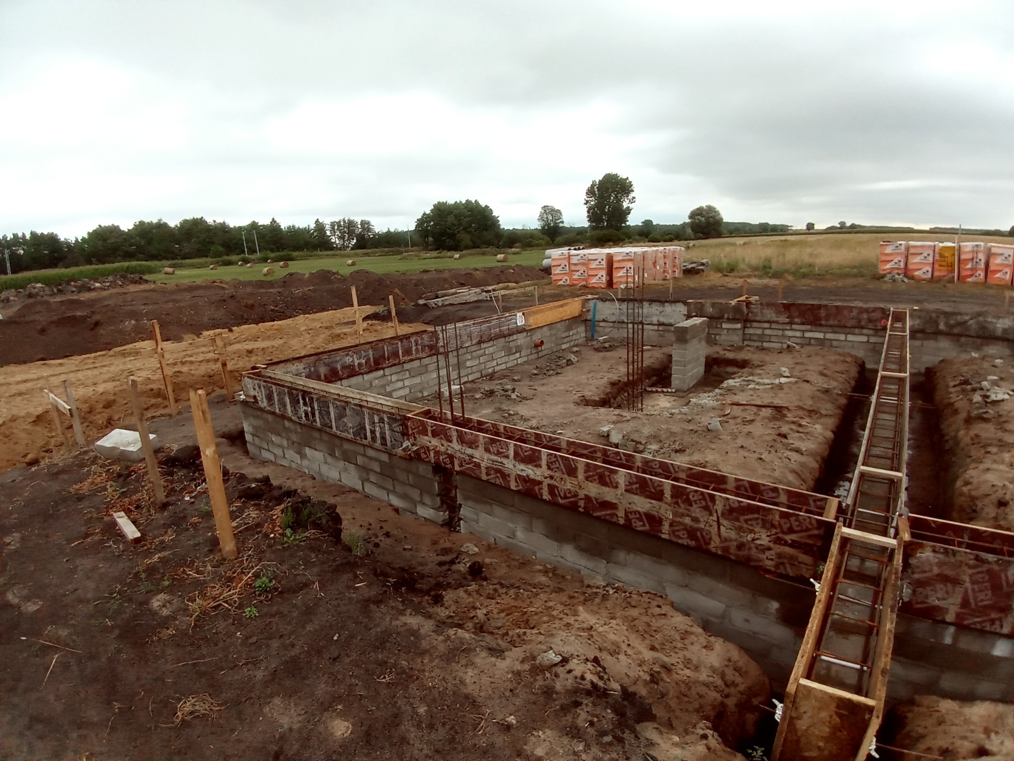 Budowa fundamentów: Wykop pod fundamenty z częściowo wylanymi ścianami fundamentowymi i szalunkami, widoczne zbrojenia, w tle palety z bloczkami budowlanymi na tle krajobrazu polnego.