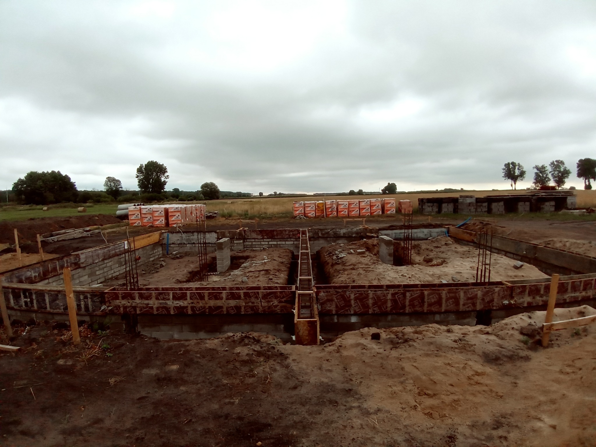 Budowa fundamentów: widok na wykop z szalunkami, stalowe pręty zbrojeniowe, bloczki fundamentowe oraz palety z cegłami w tle, na tle pochmurnego nieba i pola.