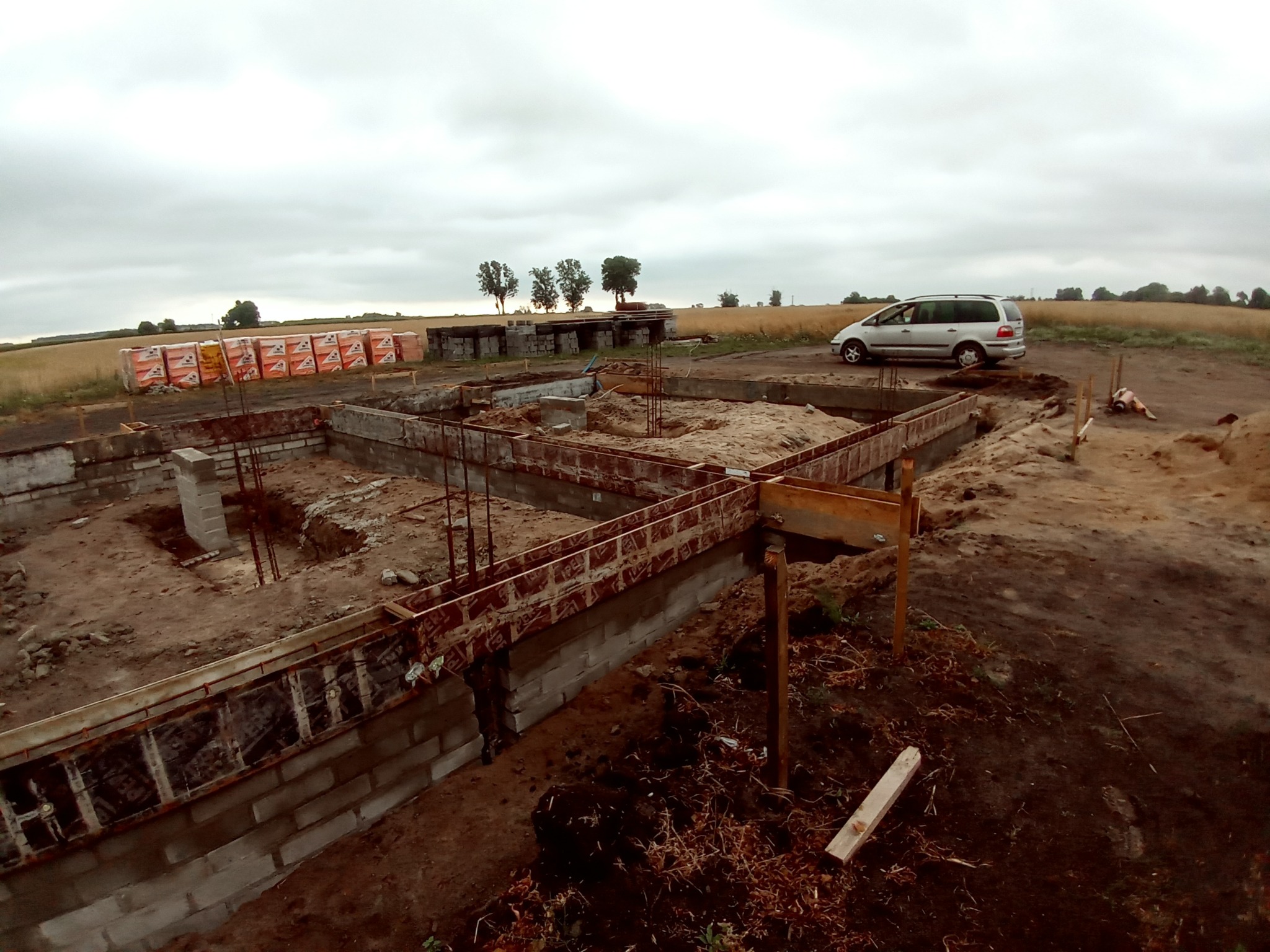 Wykopany fundament z częściowo wylaną betonową konstrukcją szalunkową, widoczne zbrojenia i zapasy cegieł, na drugim planie samochód osobowy i pole uprawne pod pochmurnym niebem.