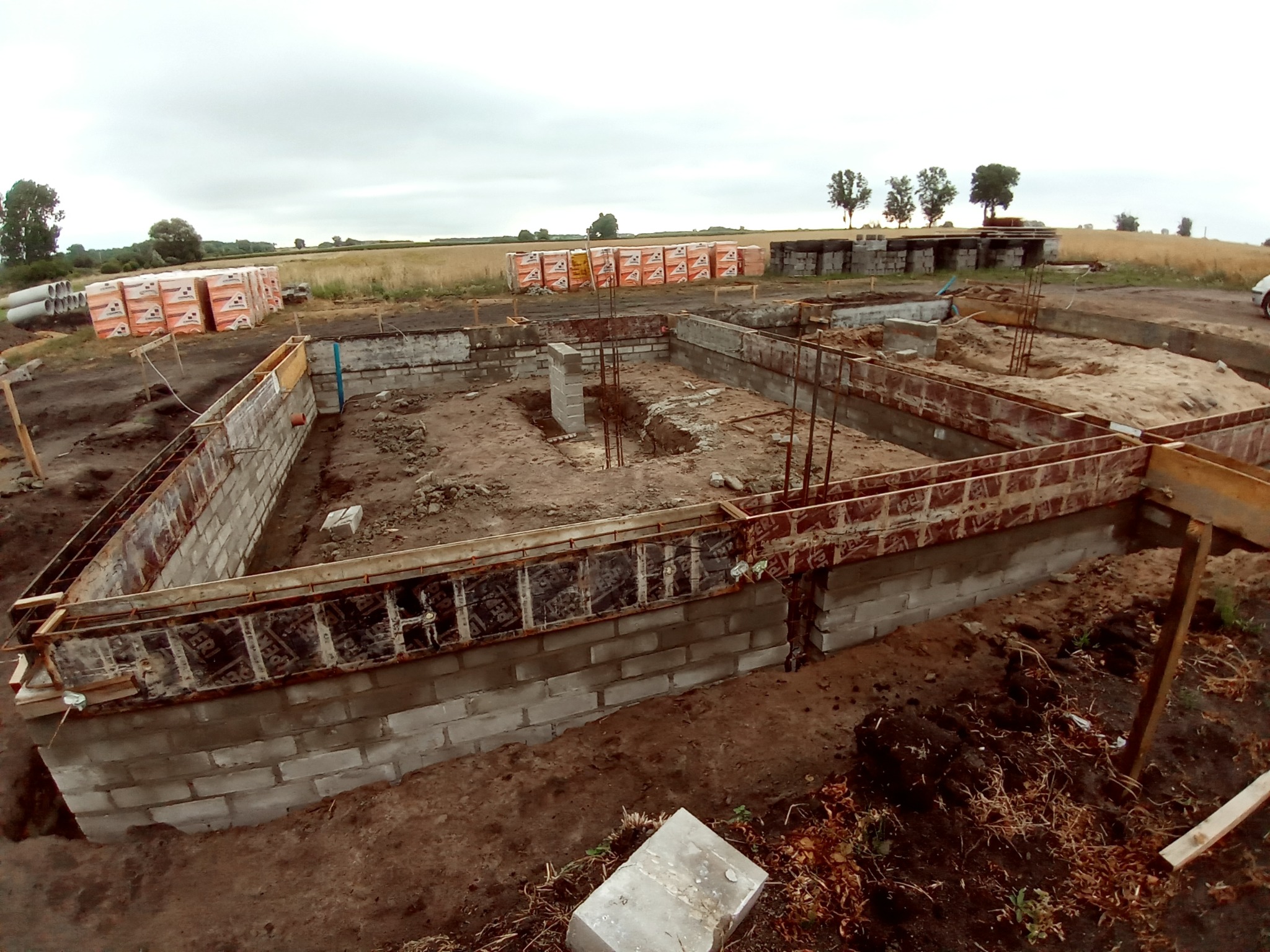 Wykop pod fundamenty z częściowo wylanymi ścianami fundamentowymi z bloczków betonowych, zabezpieczonymi folią i szalunkiem drewnianym, z wystającymi prętami zbrojeniowymi, na tle pola...