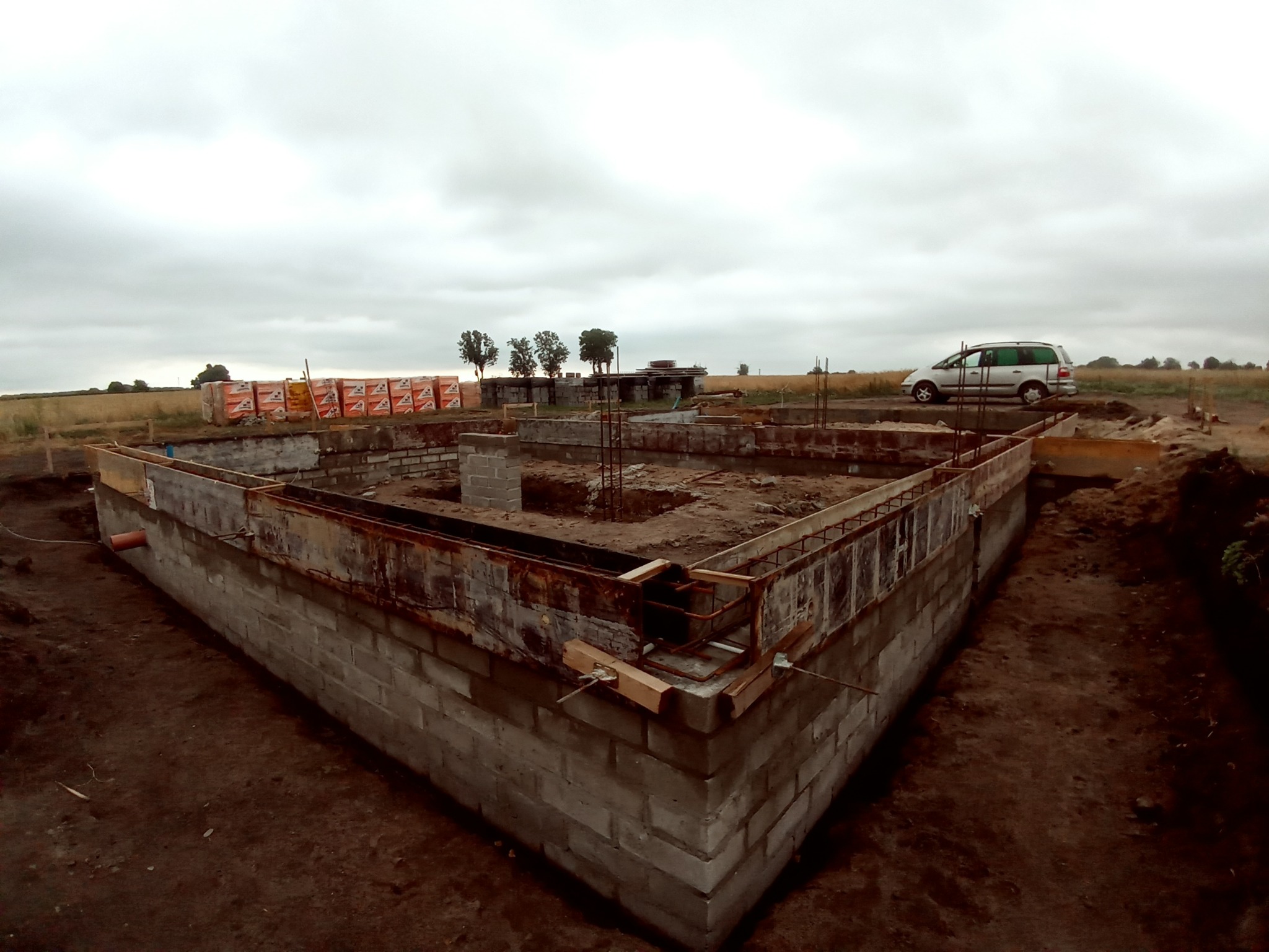 Wznoszenie fundamentów z bloczków betonowych, widoczne szalunki i zbrojenie, w tle zapas bloczków i samochód na działce budowlanej w pochmurny dzień.