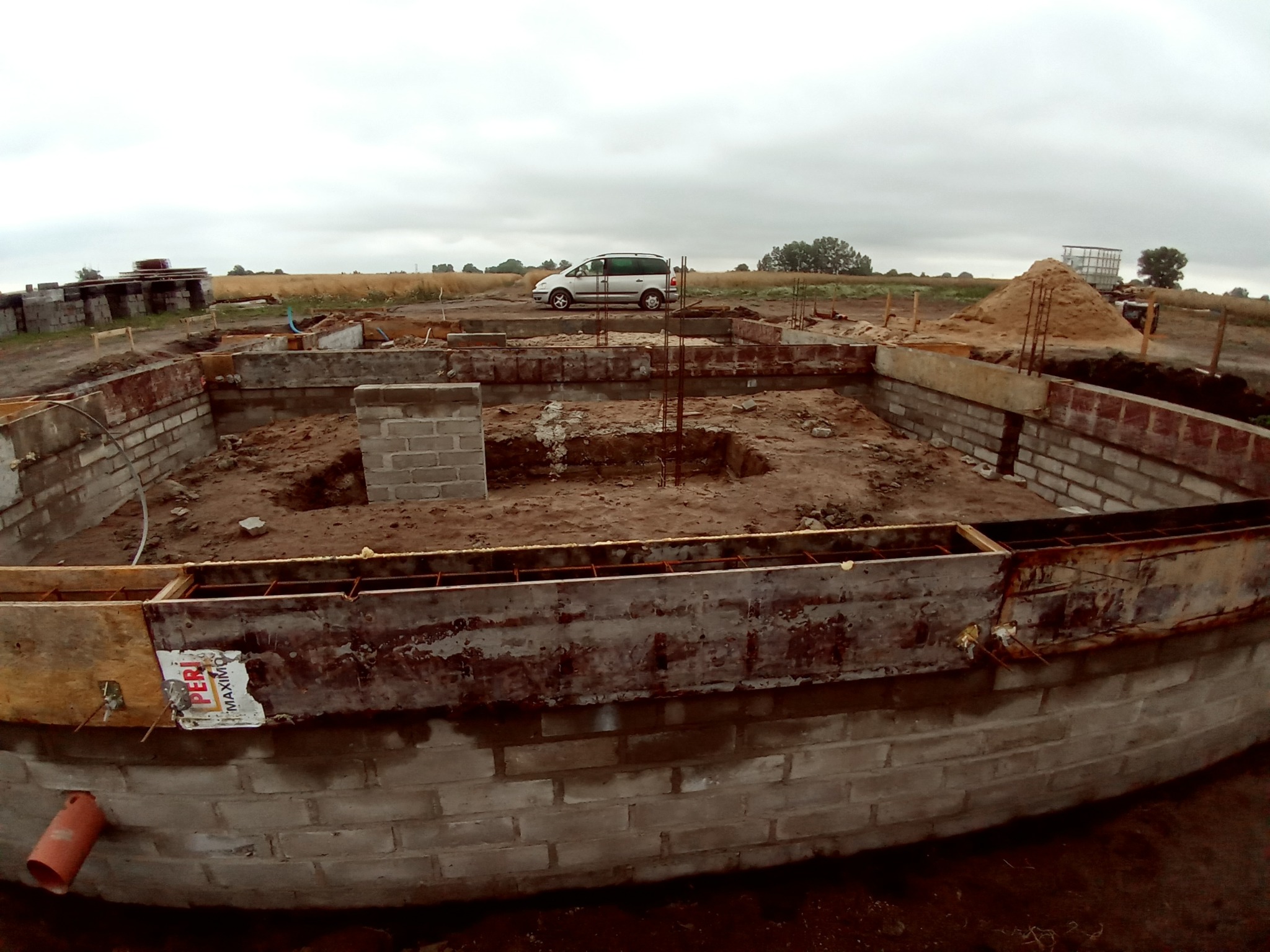 Wykop pod fundamenty z częściowo wymurowanymi ścianami z bloczków betonowych, z widocznym zbrojeniem i szalunkiem, na tle krajobrazu z samochodem dostawczym i stertą piasku.