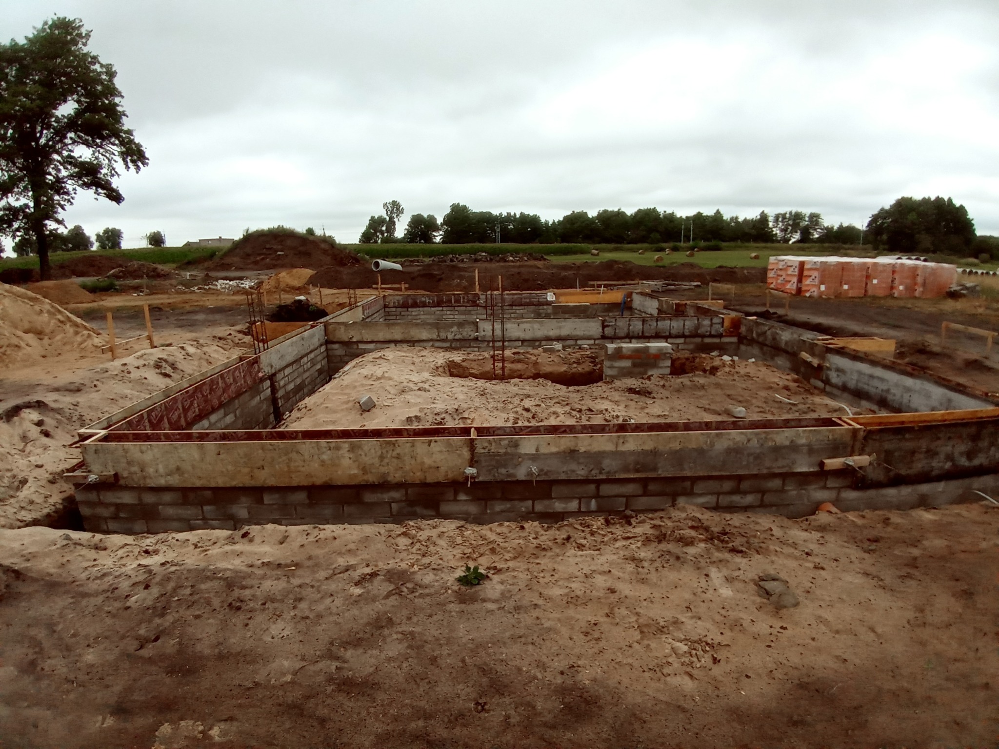 Wykopany fundament pod budowę domu z widocznymi bloczkami betonowymi i deskowaniem, w tle sterta cegieł i krajobraz polny pod pochmurnym niebem.