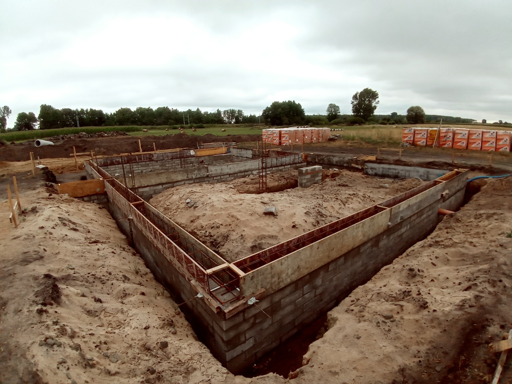 Wykop pod fundamenty domu z częściowo wylaną ławą fundamentową, zbrojenie widoczne w szalunkach, w tle palety z bloczkami betonowymi i rury kanalizacyjne.