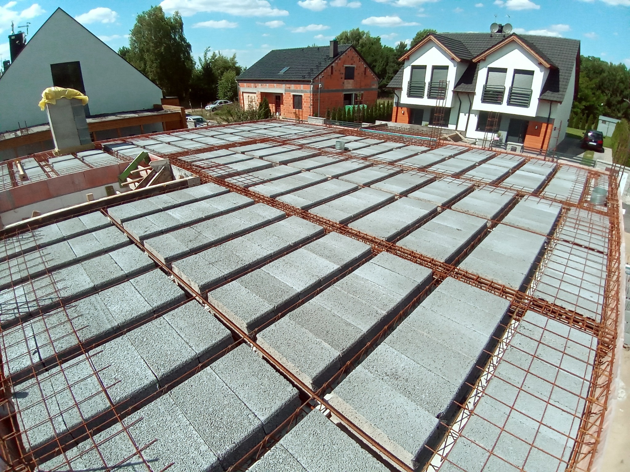 Wieniec stropowy w trakcie budowy domu murowanego, widok z góry na bloczki betonowe i zbrojenie stalowe, w tle zabudowa jednorodzinna i błękitne niebo z chmurami.