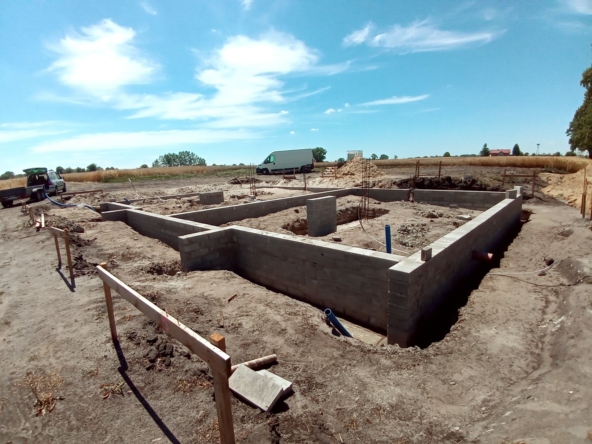 Wylane fundamenty z bloczków betonowych pod budowę domu jednorodzinnego na działce, widoczne zbrojenia, szalunki, rury instalacyjne i sprzęt budowlany w tle.